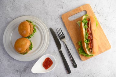 Beyaz tabakta iki hamburger ve ahşap kesim tahtasında büyük bir sosisli sandviç, domates sosu, yerde çatal ve bıçak, kopyalama alanı.