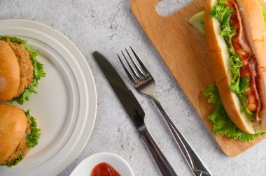 Beyaz tabakta iki hamburger ve ahşap kesim tahtasında büyük bir sosisli sandviç, domates sosu, yerde çatal ve bıçak, kopyalama alanı.