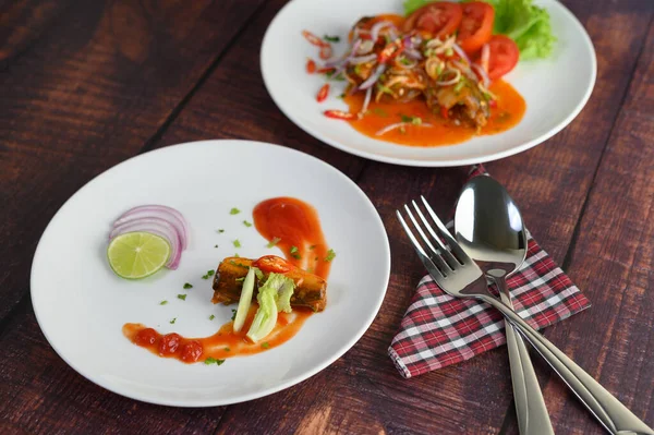 Selective focus canned sardine with tomato sauce and cooking ingredient was beautiful arrangement in white plate, stainless spoon and fork on napkin nearly on wooden table, blurred mixed sardine salad