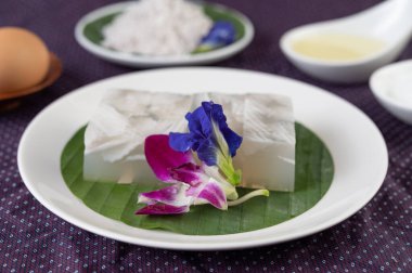 Hindistan cevizi jölesi, bir muz yaprağına konur. Kelebek bezelyesi çiçekleri ve orkidelerle bembeyaz bir tabakta..