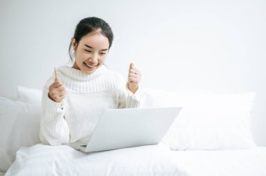 Yatakta beyaz gömlek giyen ve mutlu bir şekilde dizüstü bilgisayar oynayan bir kadın..