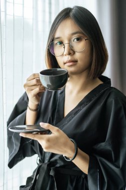 Kadın pencerenin yanında siyah bir bornoz giydi ve yatak odasında kahve içti..