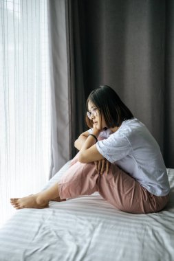 Beyaz tişörtlü bir kadın yatakta oturuyor ve dışarı bakıyor..