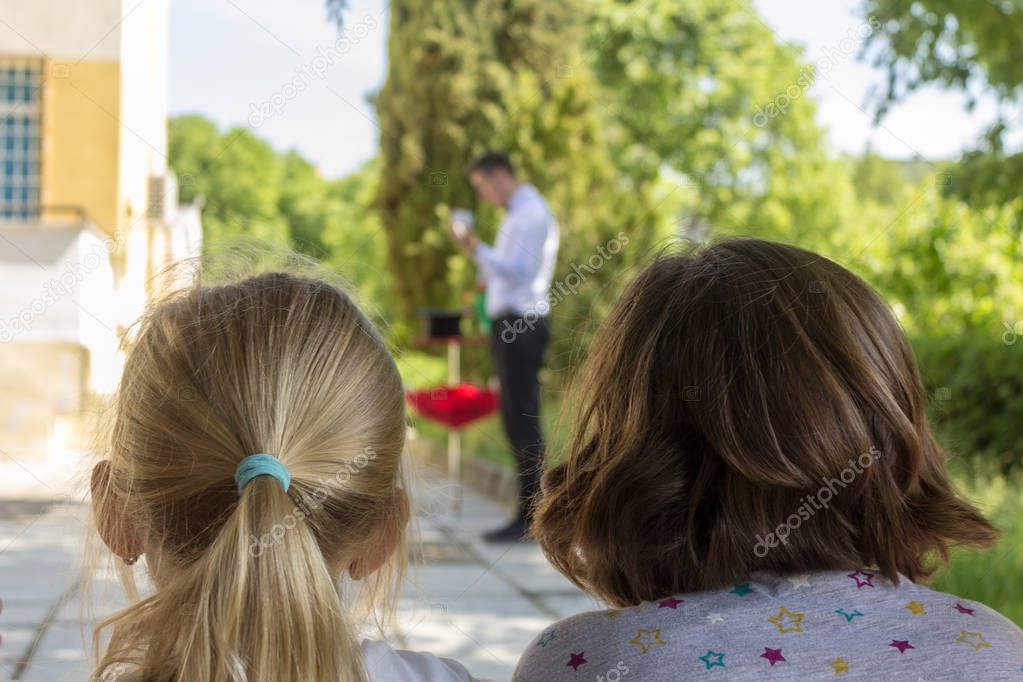 Two small girls are watching a performance of a magician through an outdoor birthday party .