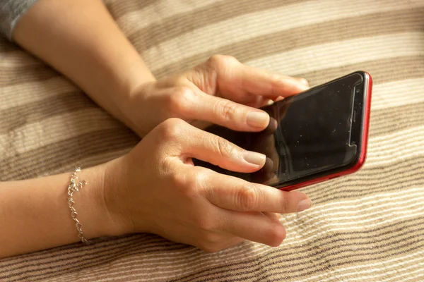 Young woman having rheumatoid arthritis takes a rest sitting on a bed, looking at her phone at home. Hands and legs are deformed. She feels pain. Selected focus.