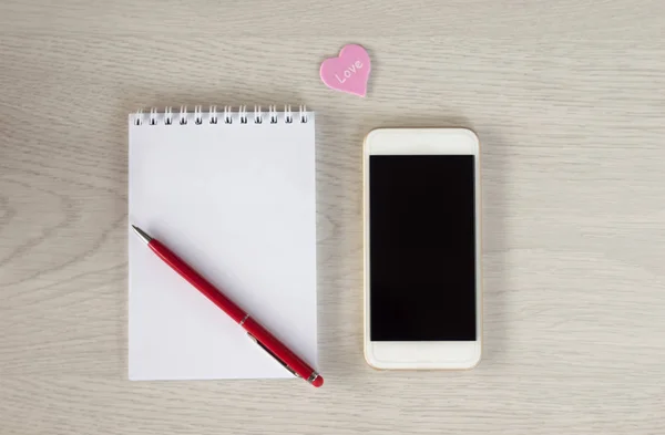 Белый телефон с блокнотом, красной ручкой и маленьким сердцем лежат на белом — стоковое фото