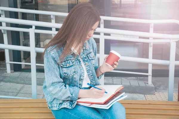 Девушка в джинсах сидит на скамейке, держит тетради, кофе — стоковое фото