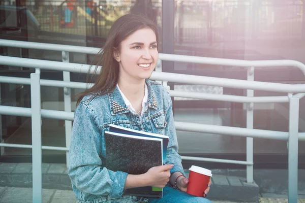 The girl in jeans clothes sits on a bench, holds notebooks and c — Stock Photo, Image