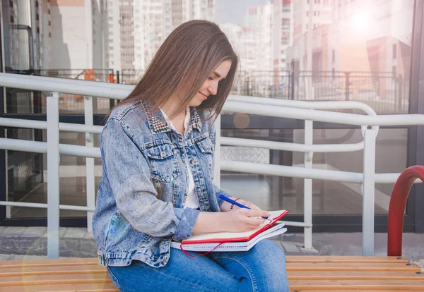 A girl in jeans clothes sits on a bench, holds notebooks and wri Royalty Free Stock Photos