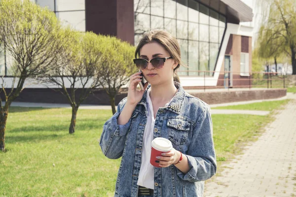 Молодая девушка в джинсовой куртке идет по улице с кофе — стоковое фото
