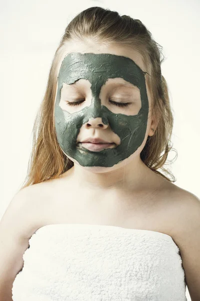 Забавная маленькая девочка с косметической маской из глины в полотенце с куском огурца, как маска, концепция красоты и здоровья, портрет в помещении крупным планом — стоковое фото