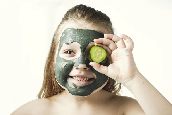 Funny little girl with a cosmetic mask of clay in a bath towel holding a piece of cucumber, like a mask, the concept of beauty and health, portrait indoors closeup