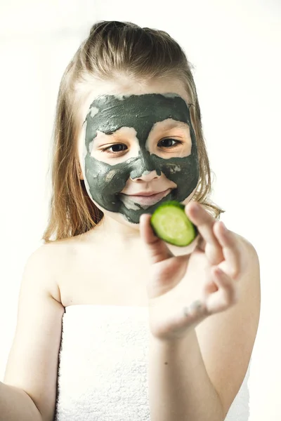 Забавная маленькая девочка с косметической маской из глины в полотенце с куском огурца, как маска, концепция красоты и здоровья, портрет в помещении крупным планом — стоковое фото