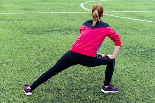 Girl in black sports leggings and a pink jacket kneads before training in an open sports arena