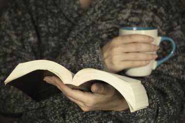 Gri örme kazak giyen genç bir kadın kitap okuyor.