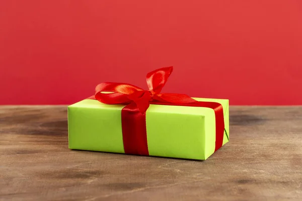 Świąteczna kompozycja. Zielone pudełko z czerwoną wstążką na drewnianym stole i czerwonym tle. Widok z boku. prezent świąteczny — Zdjęcie stockowe