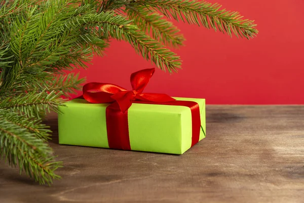 Vánoční kompozice. Zelená dárková krabice pod vánoční stromeček na dřevěném stole v blízkosti červeného pozadí. Vánoční dárek — Stock fotografie