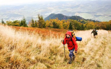 İki yürüyüşçü dağda bir patika üzerinde yürüyor. Çim kuru ve sarı. Uzaklarda dağların panoramik görünümü yoktur. Gökyüzü bulutlu.