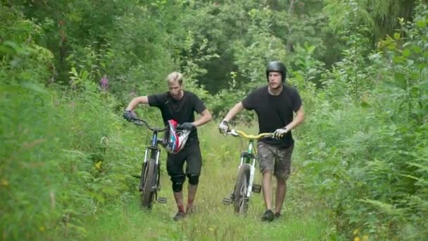 Dos ciclistas van cuesta arriba, llevan bicicletas cerca, todo alrededor de árboles de hierba y arbustos — Vídeos de Stock