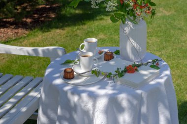 Sıcak güneşli bir bahar gününde bahçede iki kişi için masa servis. İki fincan kahve, kek, vazo, çiçek, kitap. Yanında beyaz ahşap bir bank var..