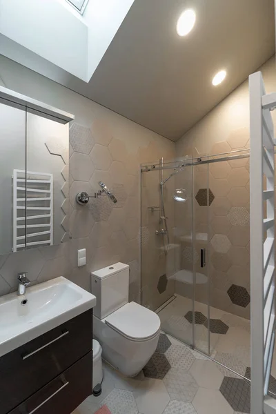 Bathroom in the attic. On the wall -  towel radiator. Shower behind transparent partition. White toilet. Cabinet with mirror on wall, below - sink with faucet on cabinet. The floors and walls are tiled. Ceiling mounted lighting