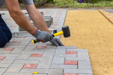 A craftsman in gloves with a rubber mallet lays concrete blocks on a gravelly sand base. Laying concrete blocks on the sidewalk clipart