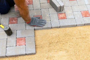 Eldiven giymiş, lastik çekiçle beton bloklar döşeyen bir işçinin elleri.