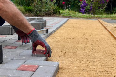 Eldivenli bir zanaatkar lastik bir çekiçle çakıllı bir kum tabanına beton bloklar yerleştirir. Eldivenli bir zanaatkar lastik bir çekiçle çakıllı bir kum tabanına beton bloklar yerleştirir. Kaldırıma beton bloklar döşemek