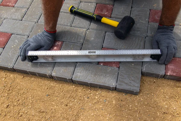 A craftsman in gloves with a rubber mallet lays concrete blocks on a gravelly sand base. Laying concrete blocks on the sidewalk
