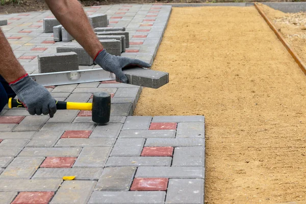 A craftsman in gloves with a rubber mallet lays concrete blocks on a gravelly sand base. Laying concrete blocks on the sidewalk