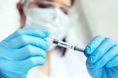 Tıbbi eldivendeki eller şırınga tutuyor. Koruyucu gözlüklü, maskeli ve eldivenli kadın doktor ellerinde şırınga tutuyor..