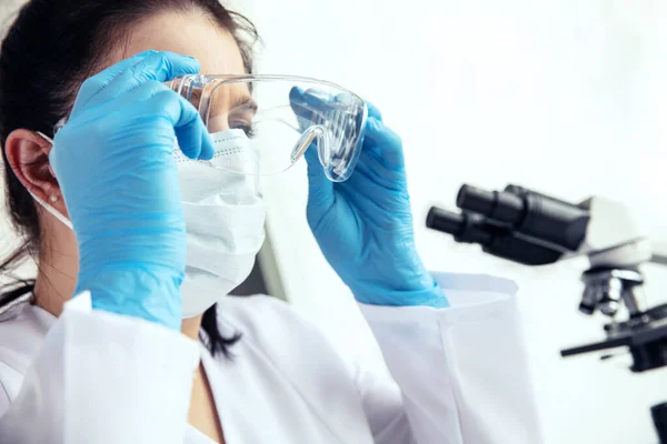 Woman scientist puts on protective glasses. Young woman doctor in medical gloves and face mask puts on protective glasses with microscope in the background.