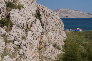 Hırvatistan, 30/06/2018: bir gökkuşağı plaj şemsiyesi çim ve Rucica kısa bir bakış, çakıllı plaja küçük yeşil bitkiler ve ünlü Pag Island çöl peyzaj kısır bir koyda yer alan