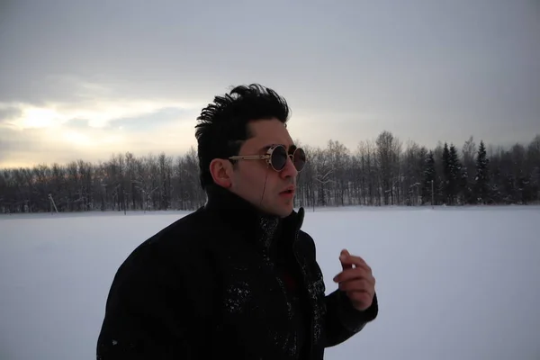 handsome young man in sunglasses stands in a field in winter. stands on a hill and looks into the distance. the majestic sky in the background