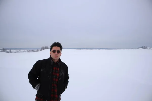 handsome young man in sunglasses stands in a field in winter. stands on a hill and looks into the distance. the majestic sky in the background