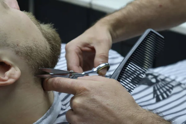 Getting perfect shape. Close-up side view with scissors and comb. Master Barber does hairstyle and styling, Bearded Man Getting Beard Haircut By Barber for brutal gay. Barber shop.