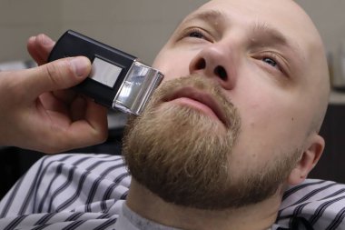 Berber sandalyeBarbershop müşteri adamın sakaltıraş. Sakal Saç Kesimi. vintage berber dükkanında elektrikli jilet ile berber tıraş sakal. Kırpma ayrıntıları. Sakal budalabir berberin kırpılmış yakın çekimi