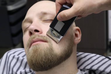 Berber sandalyeBarbershop müşteri adamın sakaltıraş. Sakal Saç Kesimi. vintage berber dükkanında elektrikli jilet ile berber tıraş sakal. Kırpma ayrıntıları. Sakal budalabir berberin kırpılmış yakın çekimi