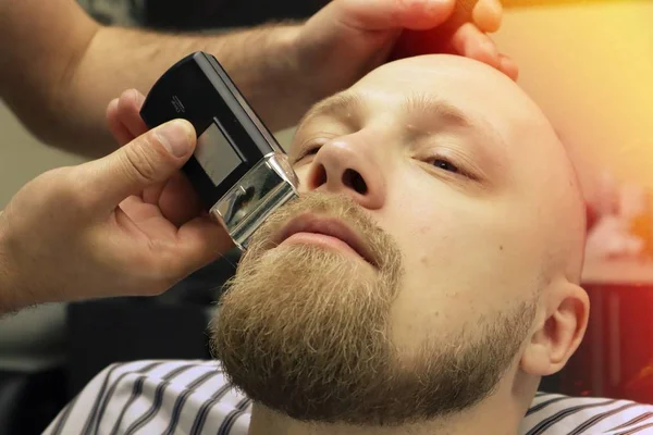 Barber shaves beard of client man on chair Barbershop. Beard Haircut. barber shaving beard with electric razor in vintage barber shop. Details of trimming. Cropped closeup of a barber trimming beard