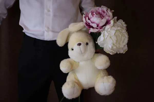 young man in a white shirt and black pants holding a toy hare and flowers. A man with  peonies and a big toy rabbit. a man in love. a gift for the woman I love