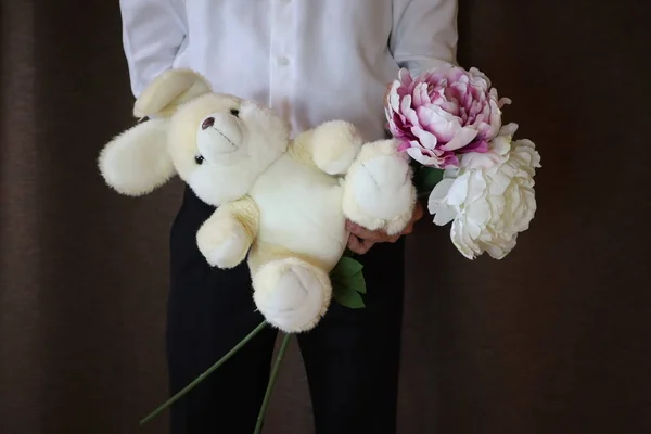 young man in a white shirt and black pants holding a toy hare and flowers. A man with  peonies and a big toy rabbit. a man in love. a gift for the woman I love