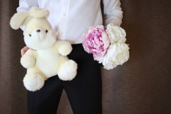 young man in a white shirt and black pants holding a toy hare and flowers. A man with  peonies and a big toy rabbit. a man in love. a gift for the woman I love