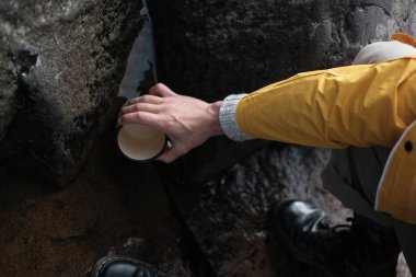 Sarı yağmurluk giyen genç bir adam elinde bir kupa tutuyor. yukarıdan görüntüleyin. yürüyüş geçmişi, seyahat. Sarı yağmurluklu ve metal kupa tutan gezgin. Rusya'da yürüyüş yapıyor hipster adam, Karelia. Atmosferik moment