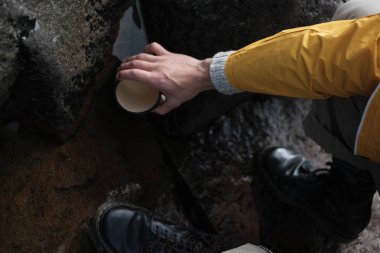 Sarı yağmurluk giyen genç bir adam elinde bir kupa tutuyor. yukarıdan görüntüleyin. yürüyüş geçmişi, seyahat. Sarı yağmurluklu ve metal kupa tutan gezgin. Rusya'da yürüyüş yapıyor hipster adam, Karelia. Atmosferik moment