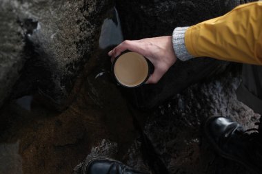 Sarı yağmurluk giyen genç bir adam elinde bir kupa tutuyor. yukarıdan görüntüleyin. yürüyüş geçmişi, seyahat. Sarı yağmurluklu ve metal kupa tutan gezgin. Rusya'da yürüyüş yapıyor hipster adam, Karelia. Atmosferik moment