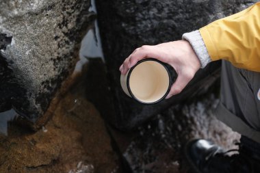 Sarı yağmurluk giyen genç bir adam elinde bir kupa tutuyor. yukarıdan görüntüleyin. yürüyüş geçmişi, seyahat. Sarı yağmurluklu ve metal kupa tutan gezgin. Rusya'da yürüyüş yapıyor hipster adam, Karelia. Atmosferik moment