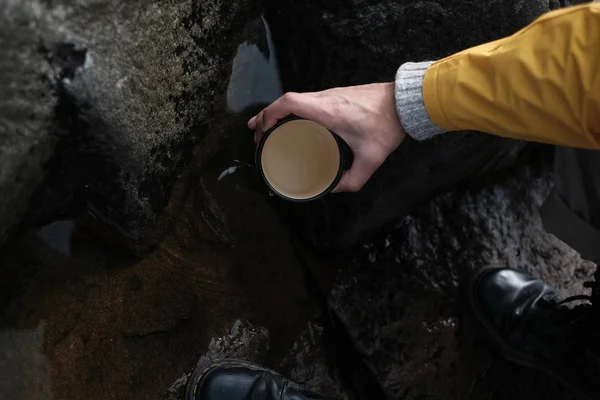 Sarı yağmurluk giyen genç bir adam elinde bir kupa tutuyor. yukarıdan görüntüleyin. yürüyüş geçmişi, seyahat. Sarı yağmurluklu ve metal kupa tutan gezgin. Rusya'da yürüyüş yapıyor hipster adam, Karelia. Atmosferik moment