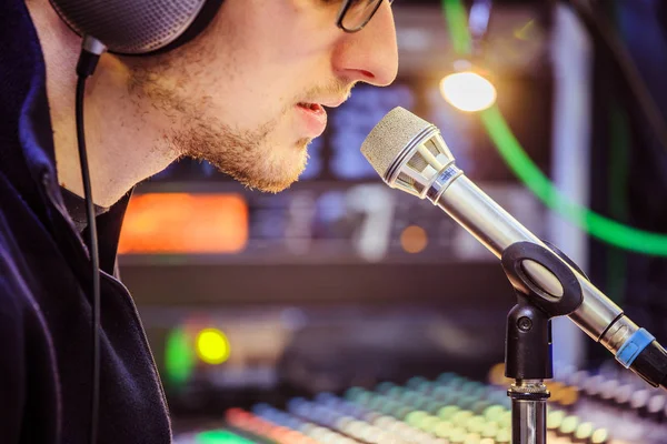 Young man is sitting in the recording studio and talks into the microphone, buttons and equipment in the blurry background