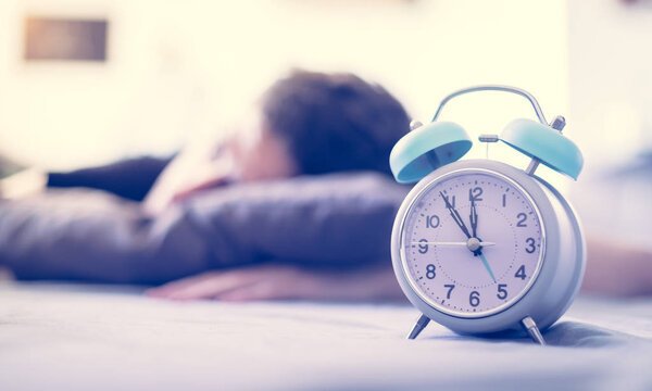 White alarm clock in the morning. Young man sleeps in the background.