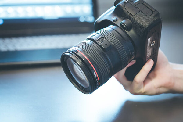 Photographer is holding a professional camera with telephoto lens in his hand, laptop in the blurry background